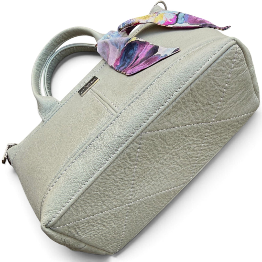 A light sage green pebbled leather handbag with a quilted bottom, dual top handles, and a colorful floral silk scarf tied to the handle, shown on a white background.