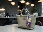 A light sage green leather handbag with top handles and a floral print neck scarf, displayed on a table in a modern cafe setting.