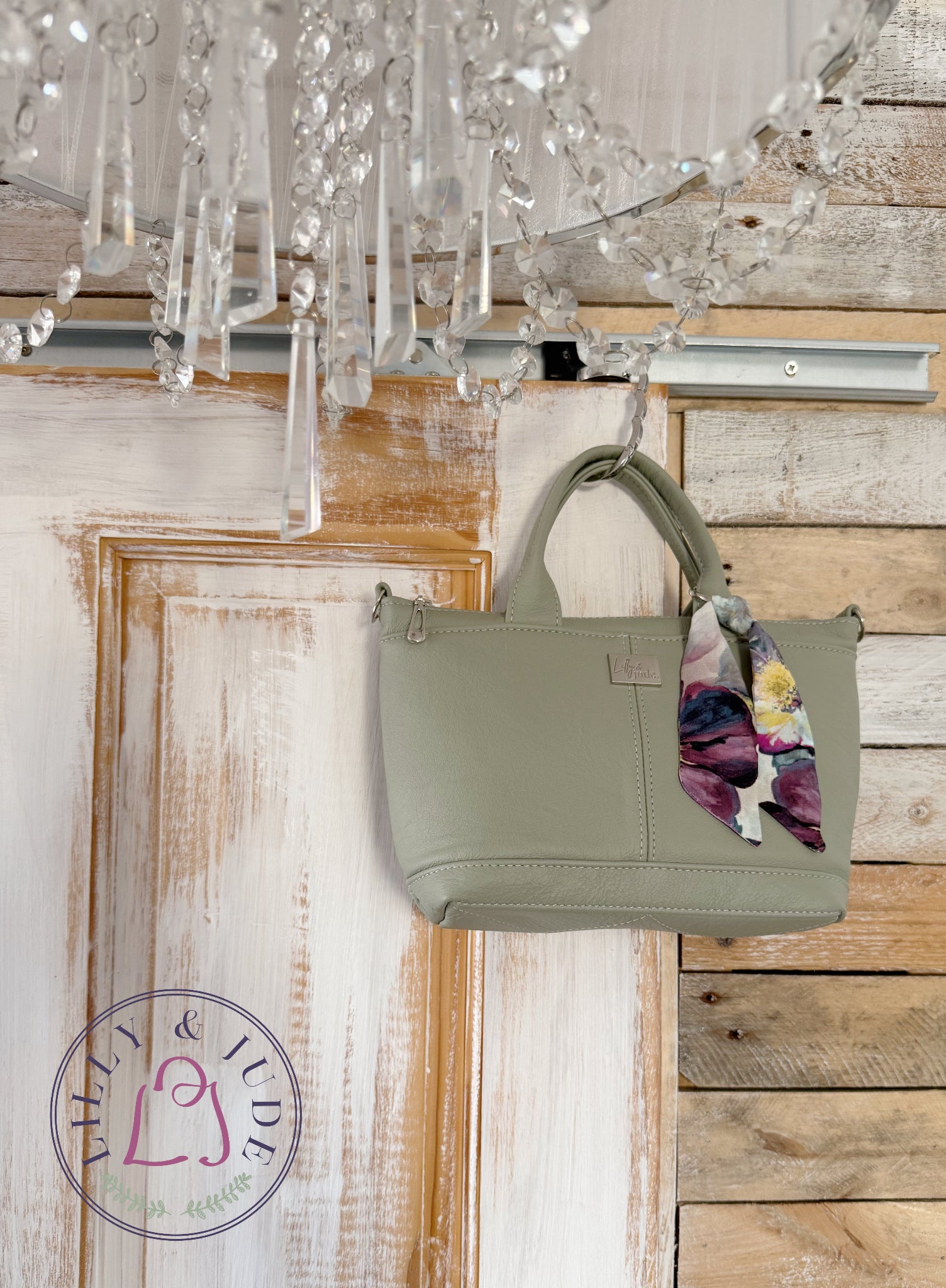 An olive green leather handbag with a floral scarf accent, hanging from a silver rod under a crystal chandelier against a rustic white and wood panelled wall.