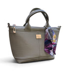 An olive green repurposed leather handbag with dual top handles, a zippered top, a small silver-tone brand plaque and a colourful floral scarf hanging from the side.
