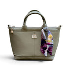 An olive green faux-leather handbag with dual top handles, a zipper opening, a small silver-tone logo plaque, and a colourful floral scarf tied to the side, shown on a white background.