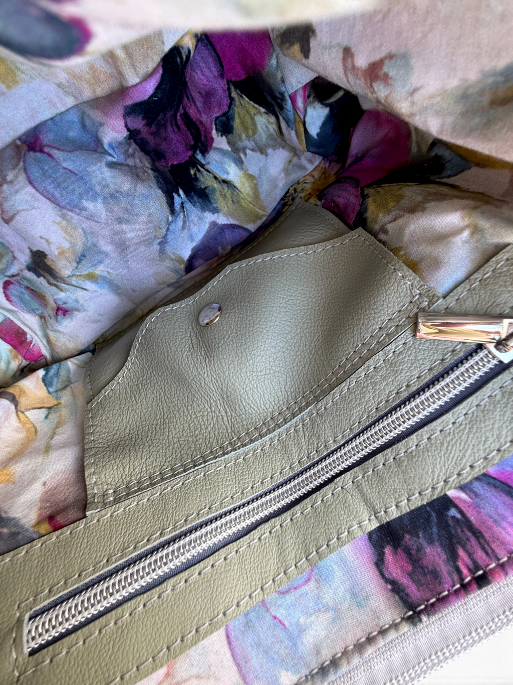 Close-up of a handbag interior showing a light olive green leather zipper pocket with a silver zipper and integrated leather purse, line with colourful floral printed fabric