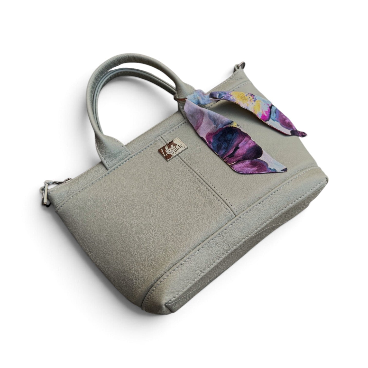 A light green gray pebbled leather handbag with dual top handles and a colourful floral scarf tied to the side, shown on a plain white background.