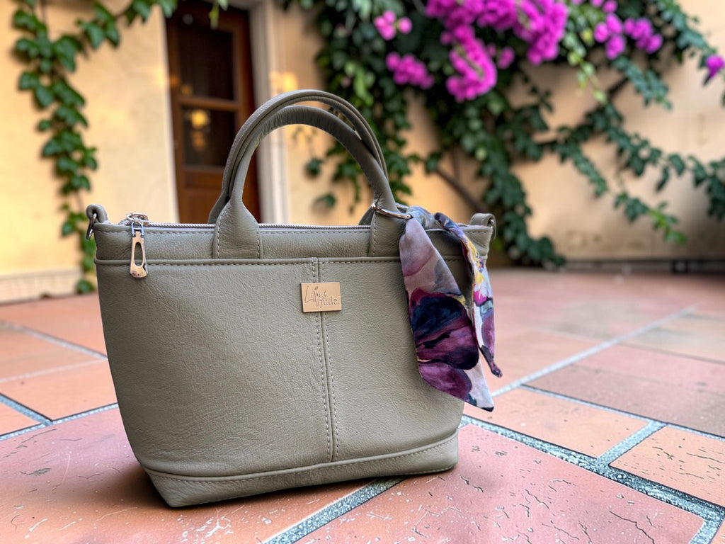 An olive green faux leather handbag with a top handle and a small gold-tone logo plaque, sitting on a terracotta tiled patio with a purple and blue floral scarf tied to its side, and a beige stucco wall with green ivy and pink flowers in the background.