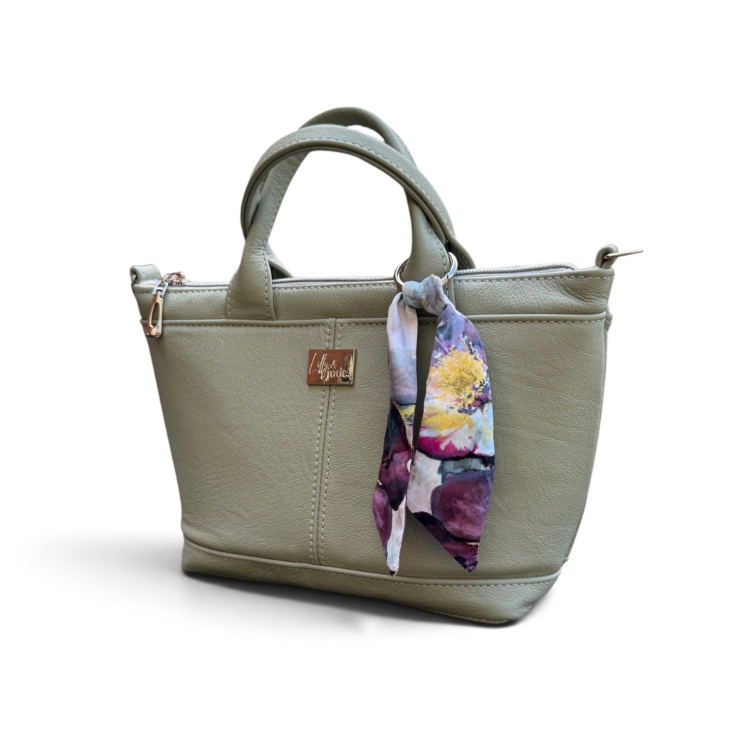 A small, olive green leather tote bag with dual top handles, a zippered top, and a colourful floral silk scarf tied to the side