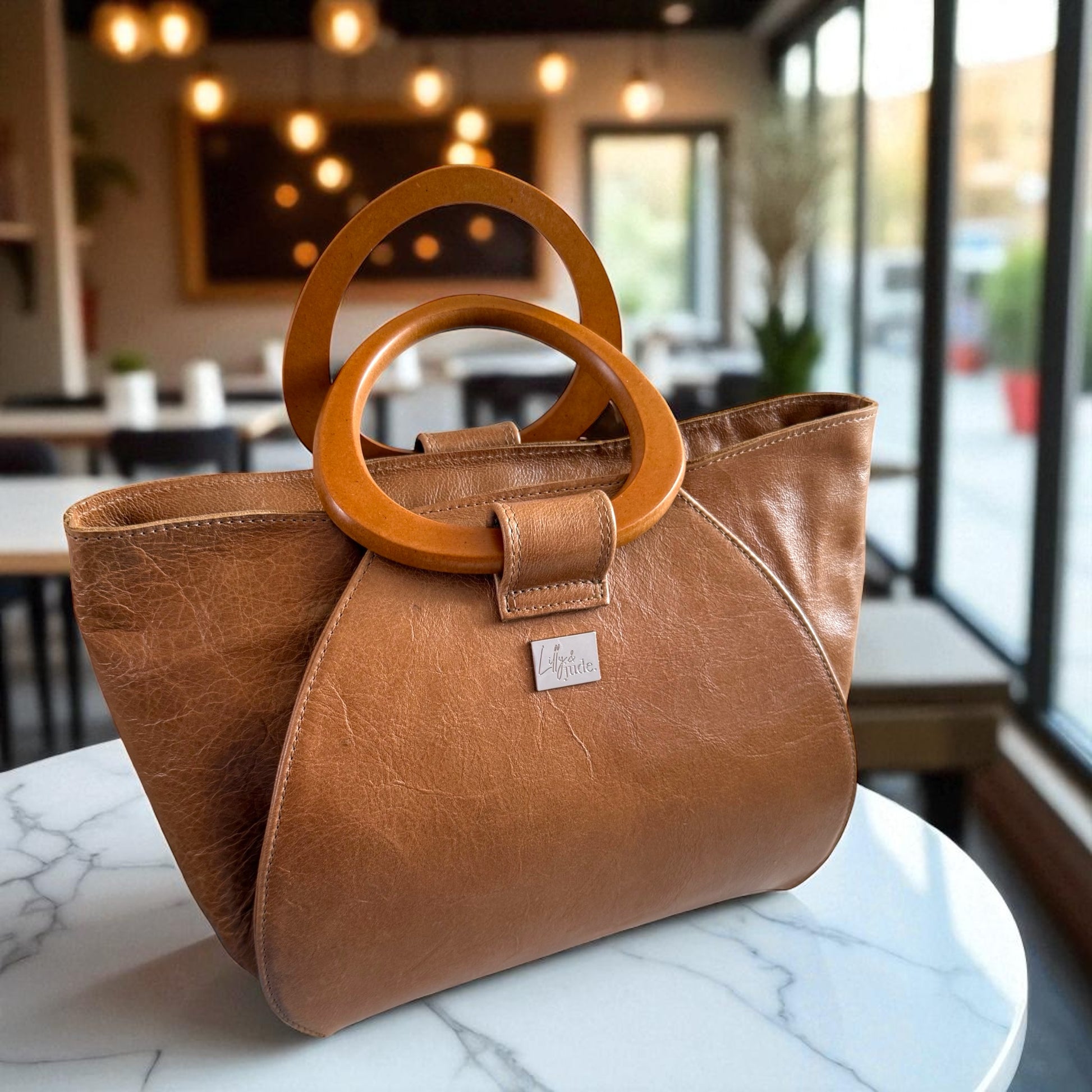 Tan leather handbag with wooden handles on a marble surface in a coffee shop setting. The bag is made from leather that has been rescued from heading to landfill