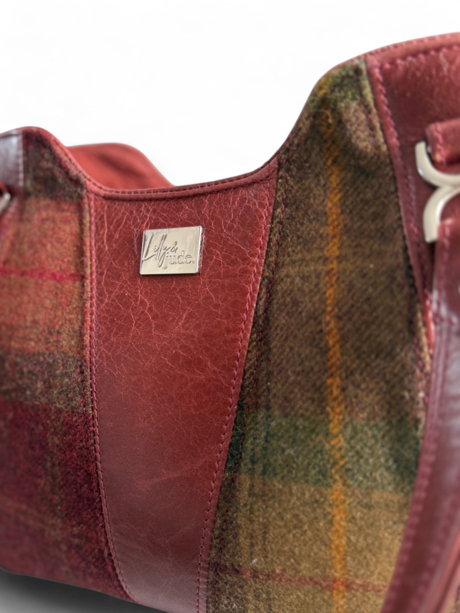 Close-up of the burgundy coloured leather Alaula handbag, with plaid pattern and leather accents, featuring silver Lilly & Jude logo label.