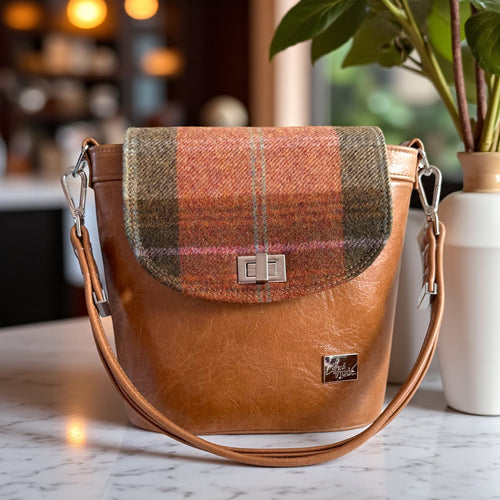 Tan brown leather bucket handbag with plaid flap  and silver twist lock on a marble surface in a coffee shop setting