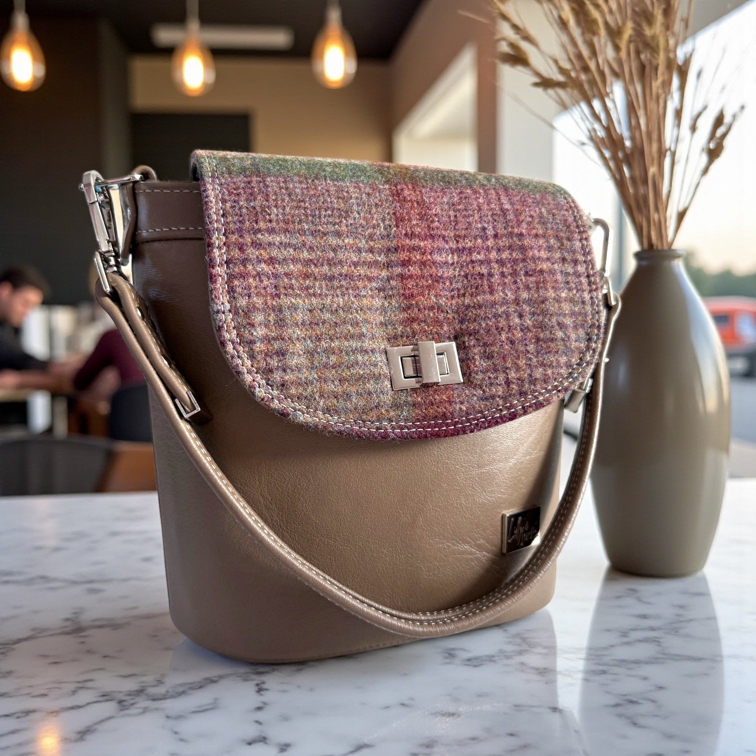 Taupe leather bucket handbag with plaid woollen tweed panels, handmade by Lilly & Jude in the UK. The bag is sat on a marble table in a cosy coffee shop
