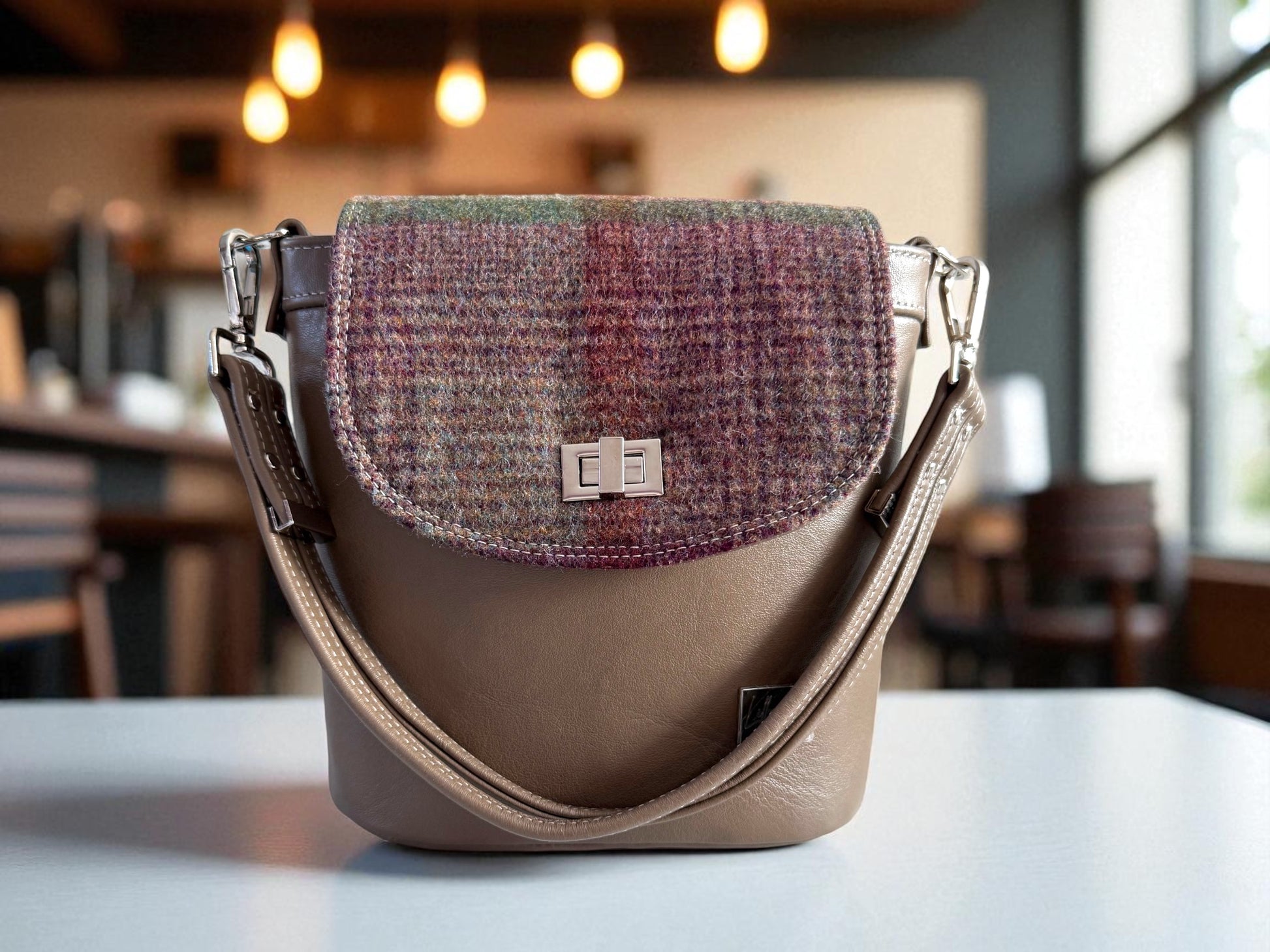 Taupe brown leather bucket bag, with heather toned plaid tweed flap on a table with a blurred indoor background