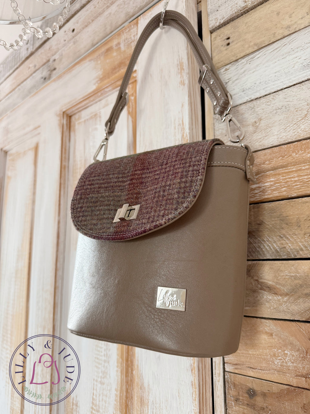 Handmade brown grey leather handbag with a woollen tweed flap hanging on a rustic wooden door. The image also contains the Lilly & Jude logo.