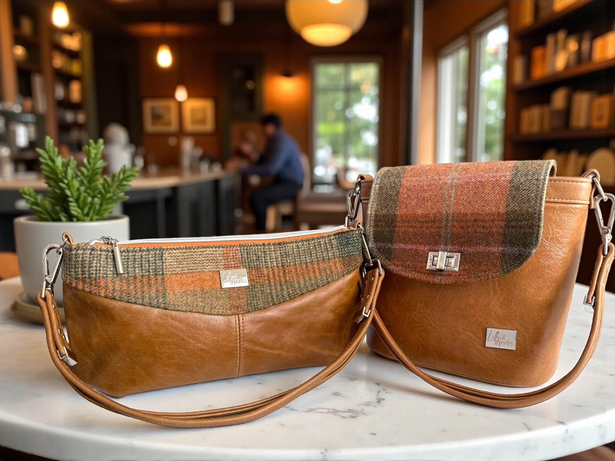 Two brown leather handbags with plaid accents on a marble surface in a cozy indoor setting. These bags are the Belle Bucket Bag and the Elland East / West Bags from the Hebden Collection at Lilly & Jude