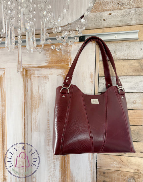 Burgundy leather Alaula handbag hanging on a rustic wooden wall with a chandelier above.
