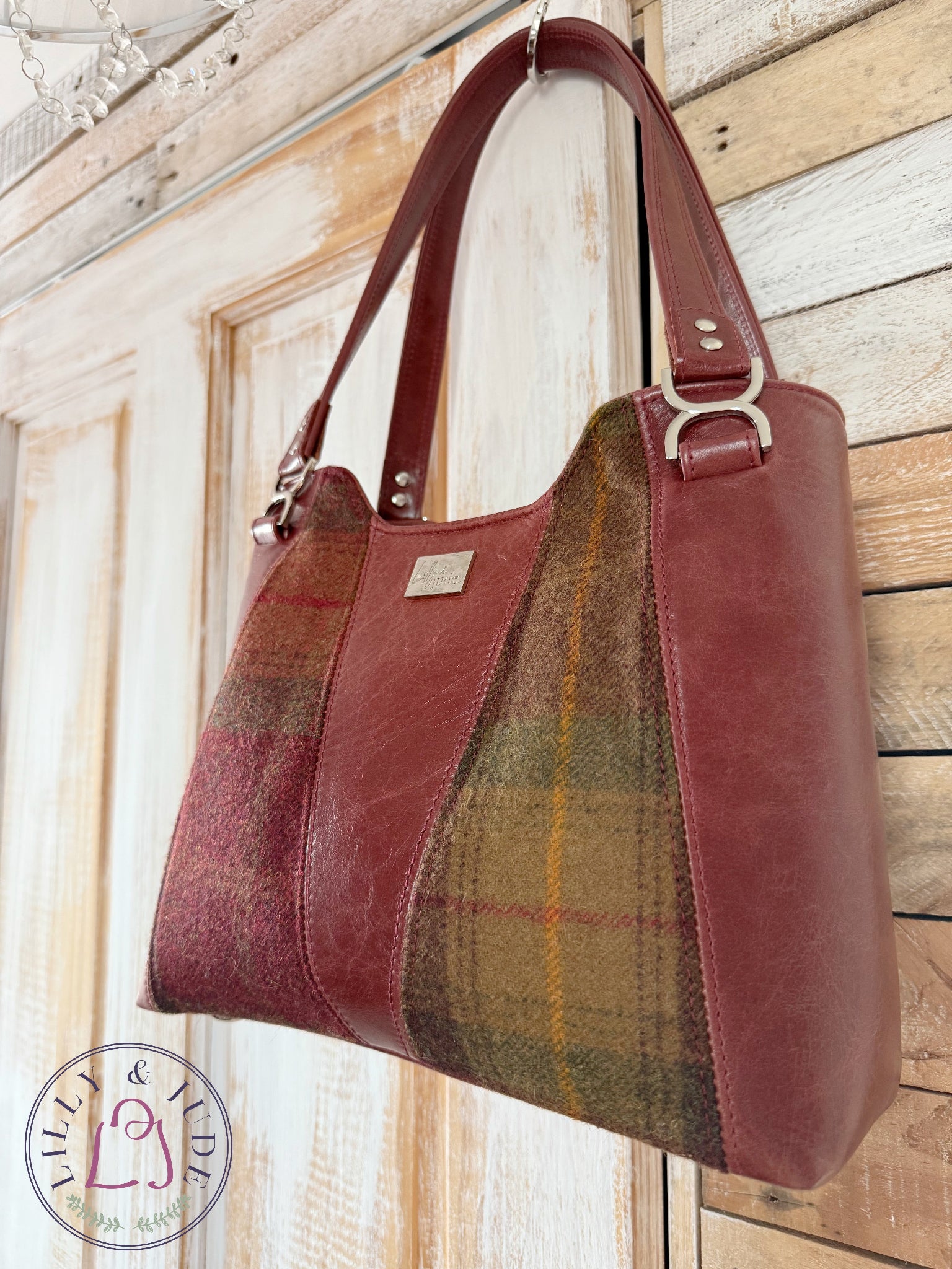 Burgundy rescued leather handbag with matching plaid patterned tweed panels, hanging from the corner of a rustic looking wooden door. 