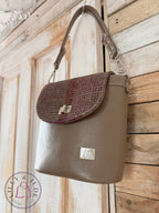 Handmade taupe leather handbag with a woollen tweed flap hanging on a rustic wooden door. The image also contains the Lilly & Jude logo.