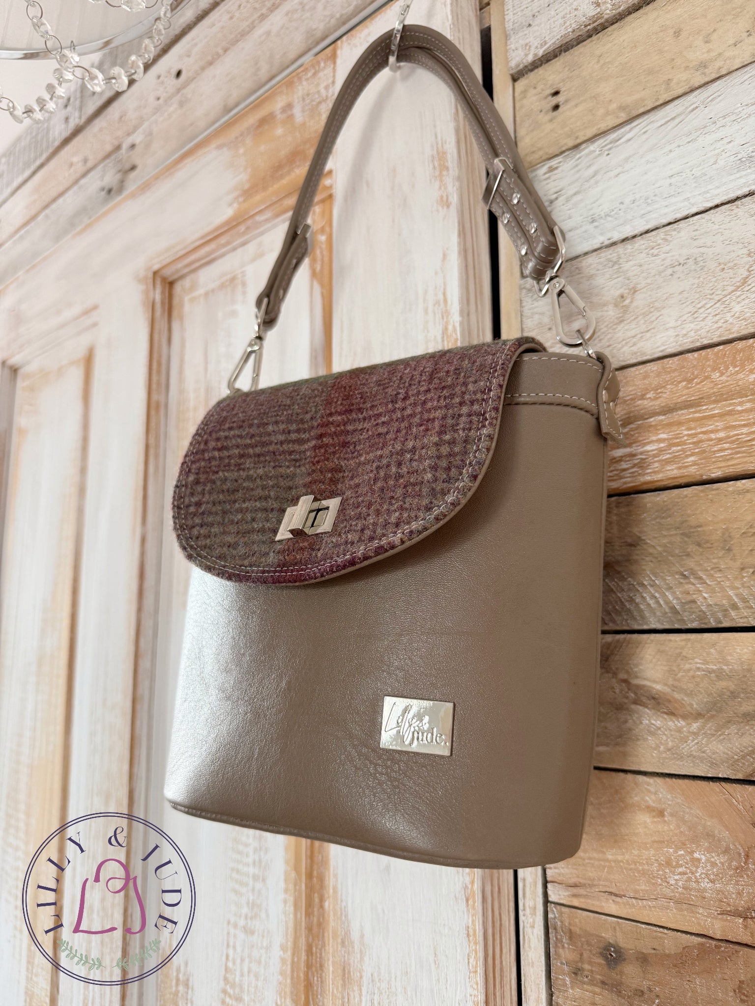Handmade taupe leather handbag with a woollen tweed flap hanging on a rustic wooden door. The image also contains the Lilly & Jude logo.