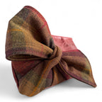 Close up view of the pllaid tweed bow with a knot on the front of a burgundy leather clutch bag on a white background