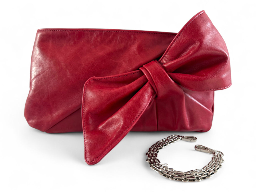 Red leather clutch with a large bow and a silver bracelet style removable chain strap on a white background. 