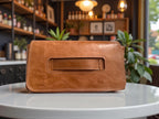 Tan leather Bronte clutch bag with integrated hand strap, sat on a table in a coffee shop