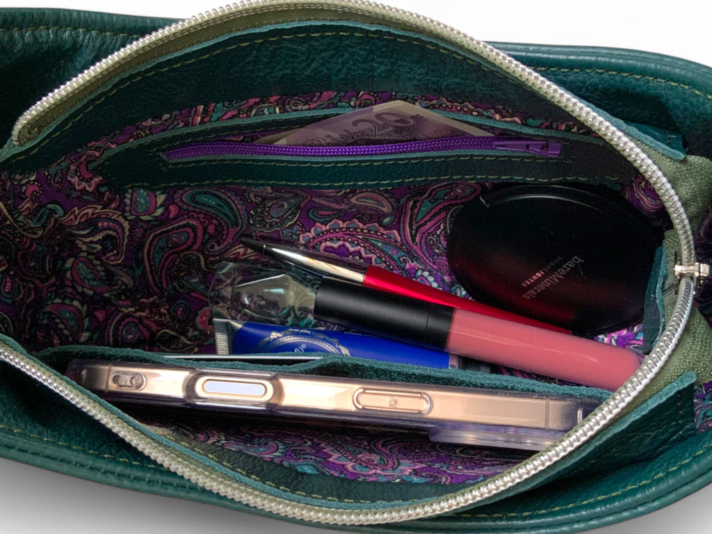 Open teal green handbag with purple paisley patterned lining, showing contents including a phone and makeup.