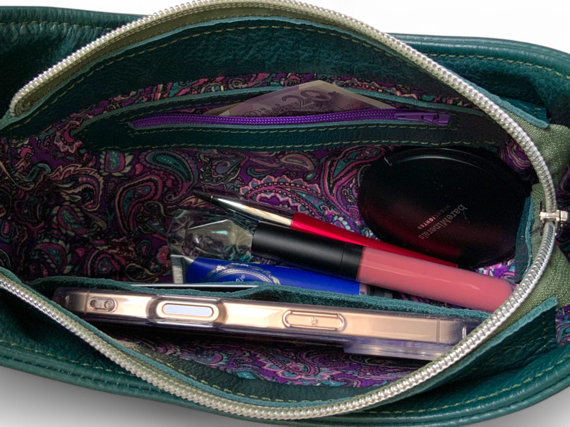 Open teal green handbag with purple paisley patterned lining, showing contents including a phone and makeup.