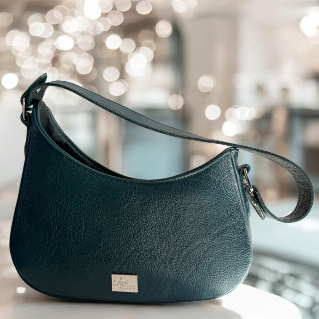 Handcrafted, dark turquoise leather handbag with silver Lilly & Jude logo, sitting on a white table with bokeh lighting in the background