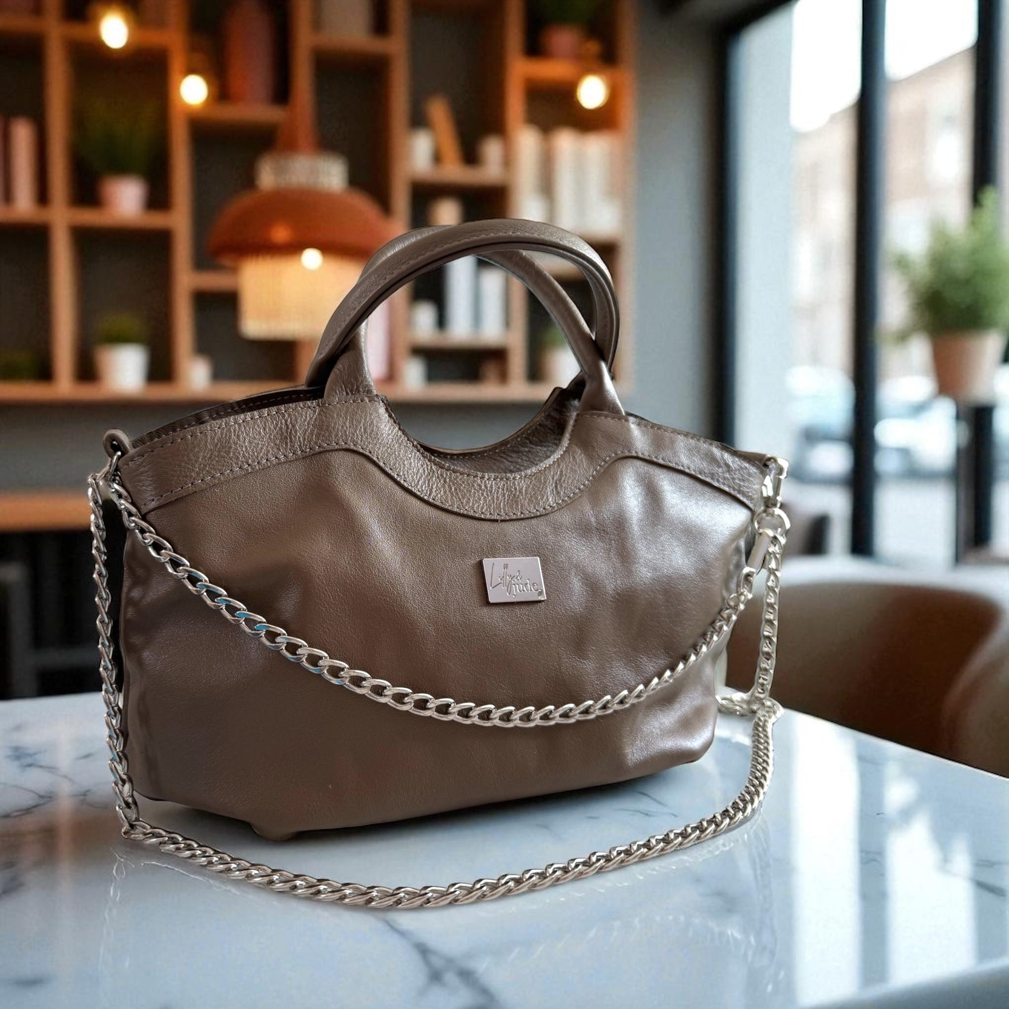 Slouchy, taupe leather handbag, with a silver chain sat on a marble table in a coffee shop setting
