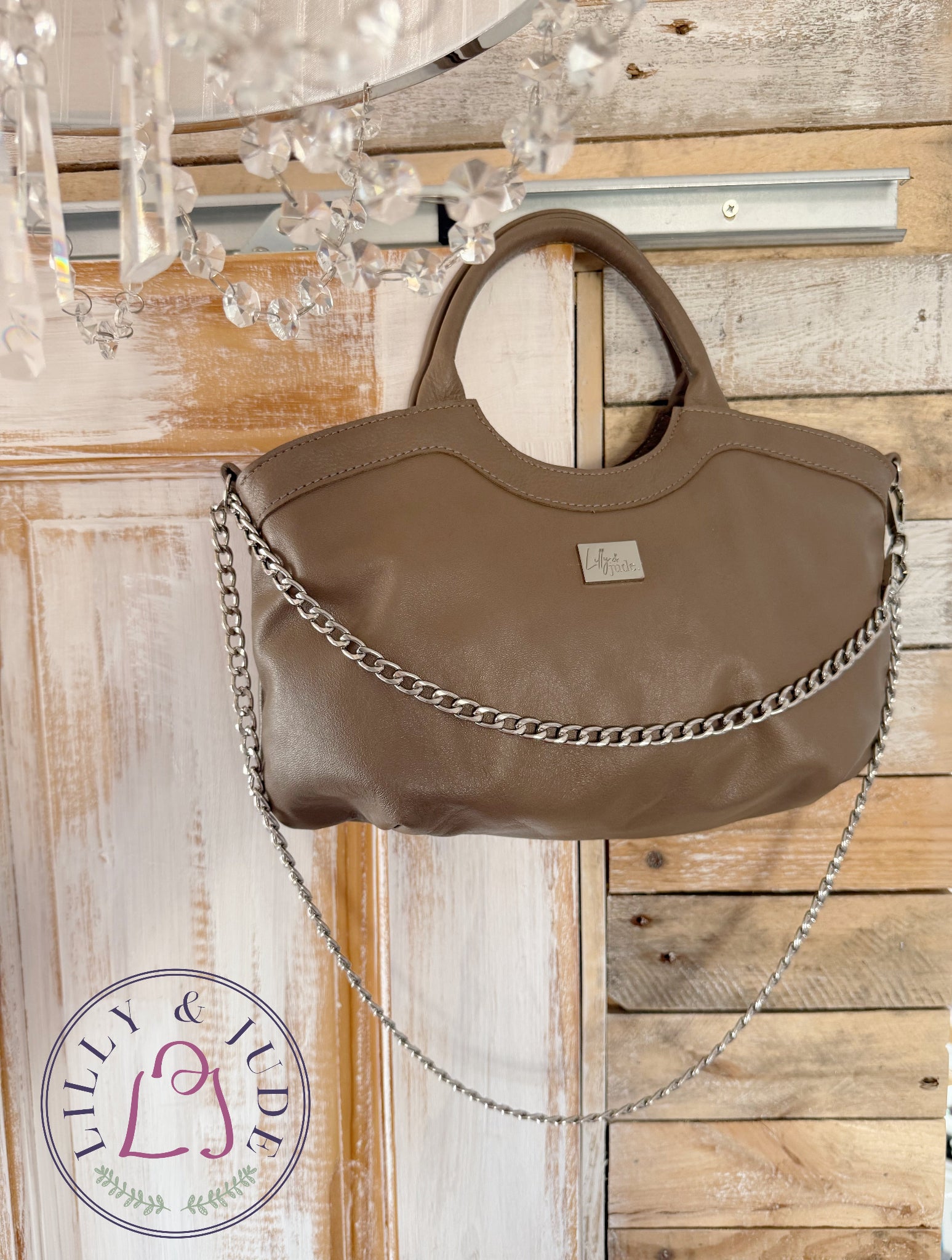 Taupe leather handbag, hanging from the top corner of a wooden door, with a chandelier in the foreground. 