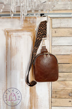 Handmade, brown leather sling bag with a patterned strap hanging from a wooden door