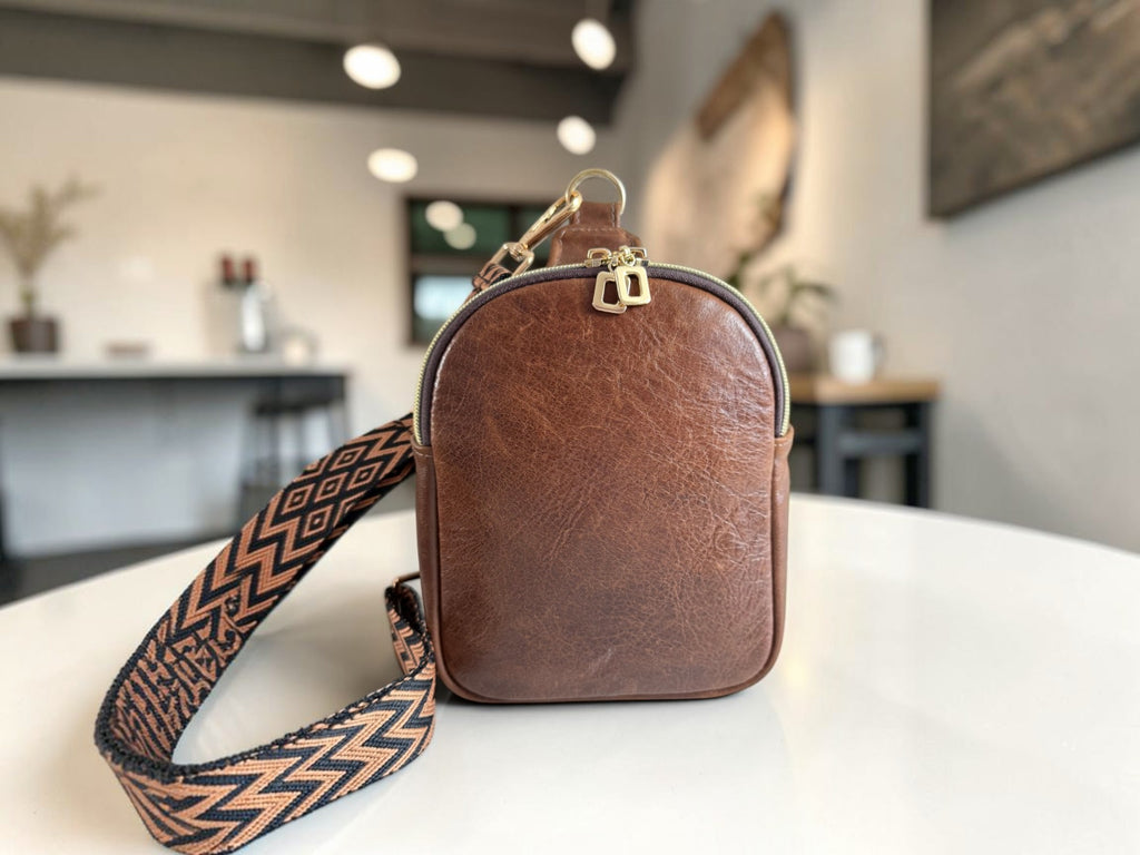 Small, brown leather crossbody sling bag with a patterned strap on a white surface against a blurred coffee shop background
