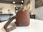 Small, brown leather crossbody sling bag with a patterned strap on a white surface against a blurred coffee shop background