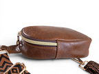 View of the side and top of the Maven crossbody  sling bag in brown leather with gold zipper lying on a white background