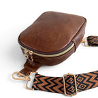 A small, brown leather sling bag with a patterned strap, lying on a white background