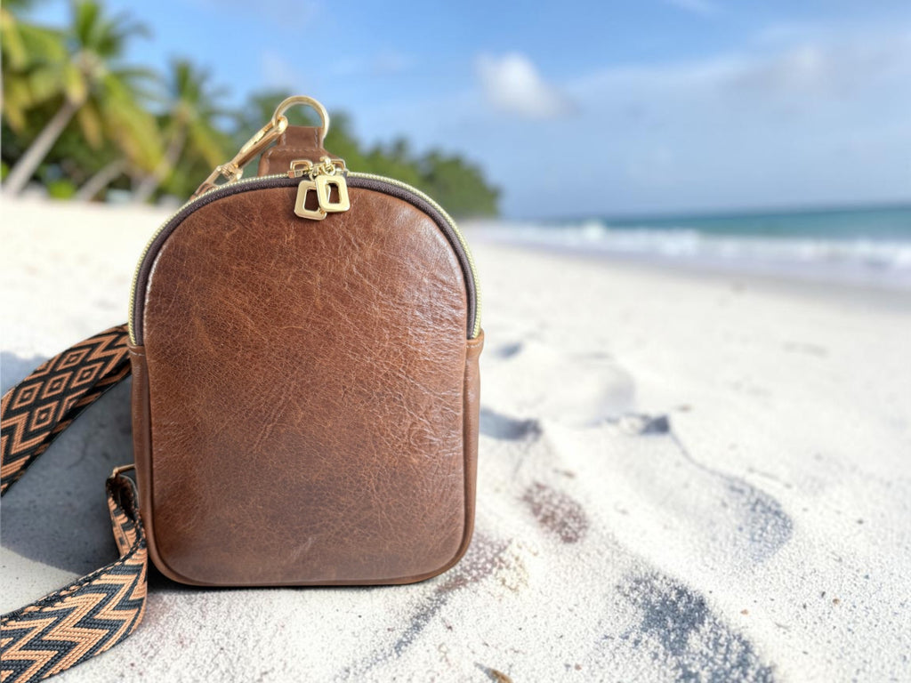 Compact, brown leather sling bag with a woven strap on a white sand beach