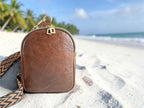 Compact, brown leather sling bag with a woven strap on a white sand beach