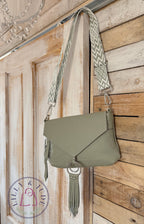 Pale sage green leather crossbody bag, with tassels and matching chevron patterned adjustable strap, hanging on a rustic looking wooden door.