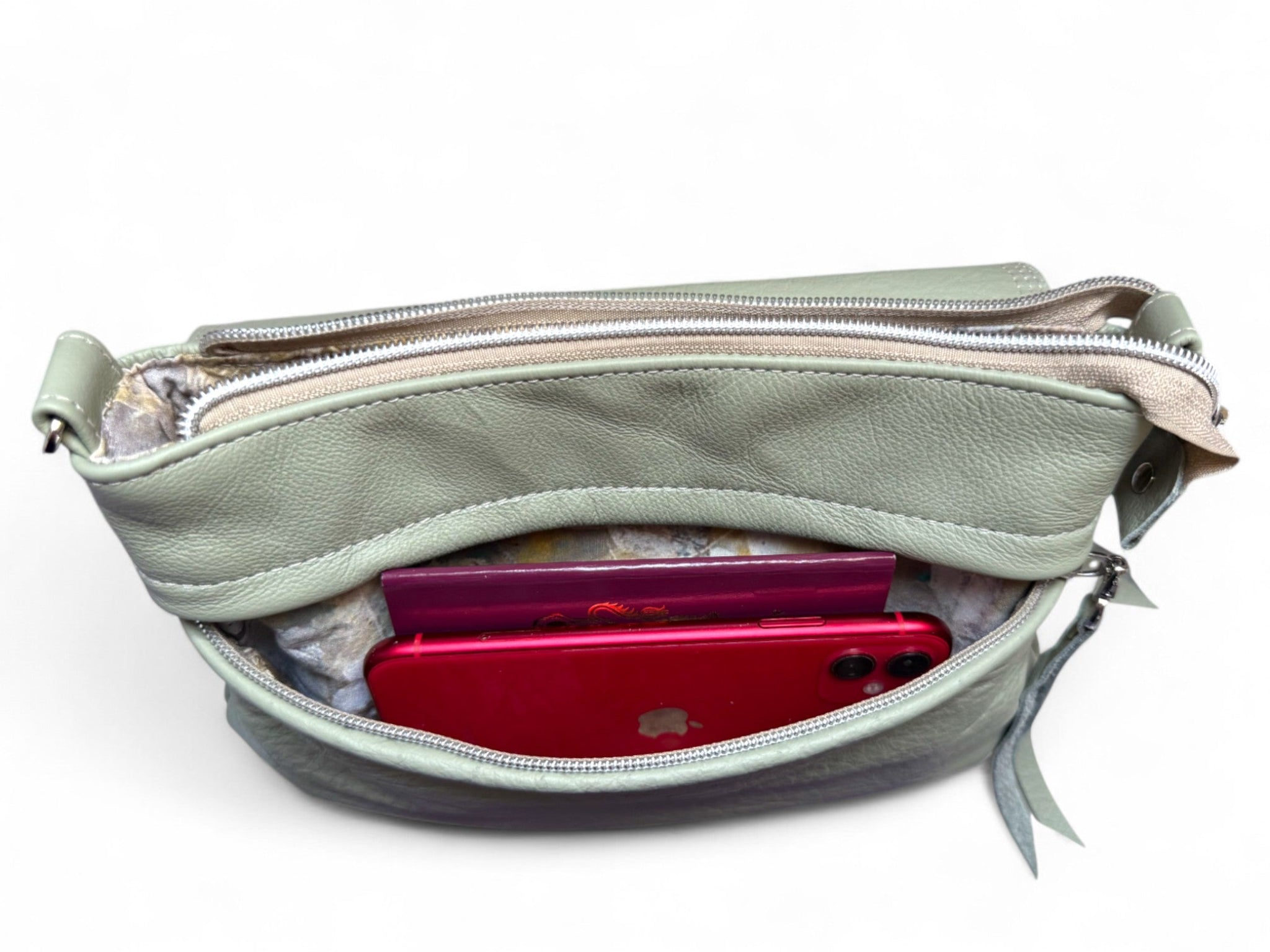 Light olive green leather bag against a white background, showing the open front pocket, with a passport and mobile phone inside.