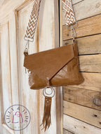 Olivia crossbody bag is tan brown leather with a woven crossbody strap and decorative, tasselled ring hanging on a wooden door. 