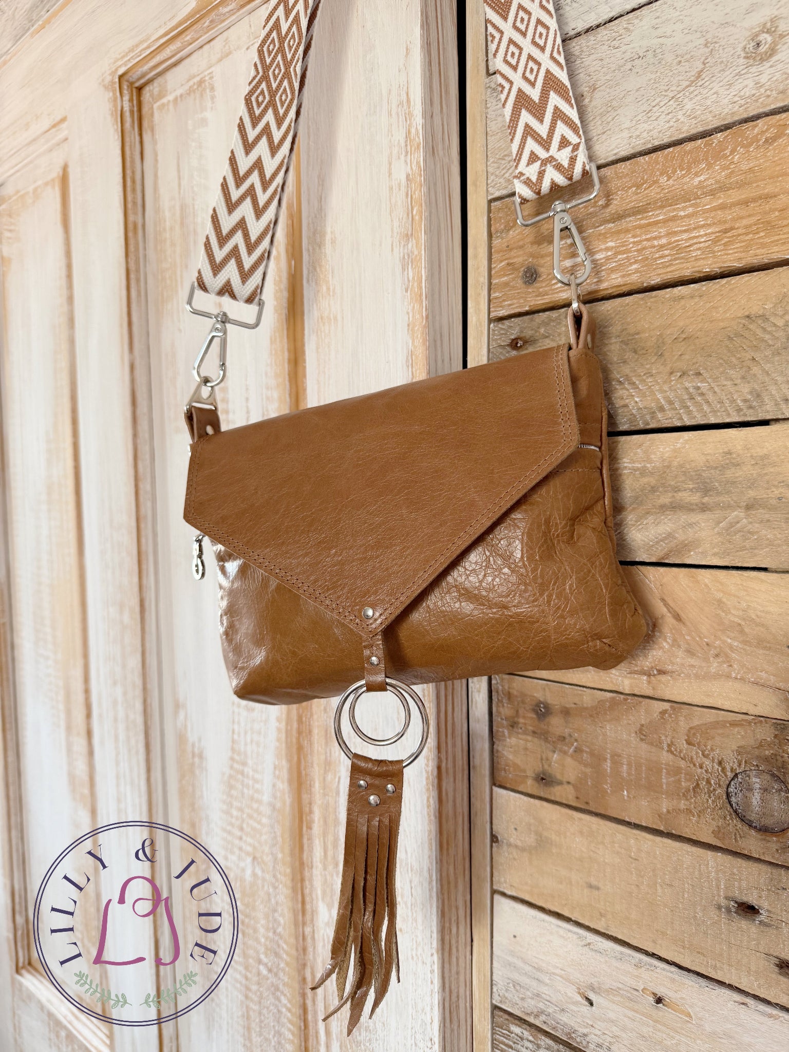 Olivia crossbody bag is tan brown leather with a woven crossbody strap and decorative, tasselled ring hanging on a wooden door. 