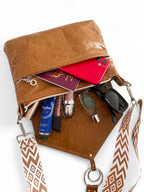tan brown leather bag, lying down on a white background, with the contents, such as cosmetics, perfume and sunglasses spilling out.