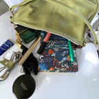 Open, soft green leather slouchy handbag with contents spilled on a white surface, including a book and makeup items.