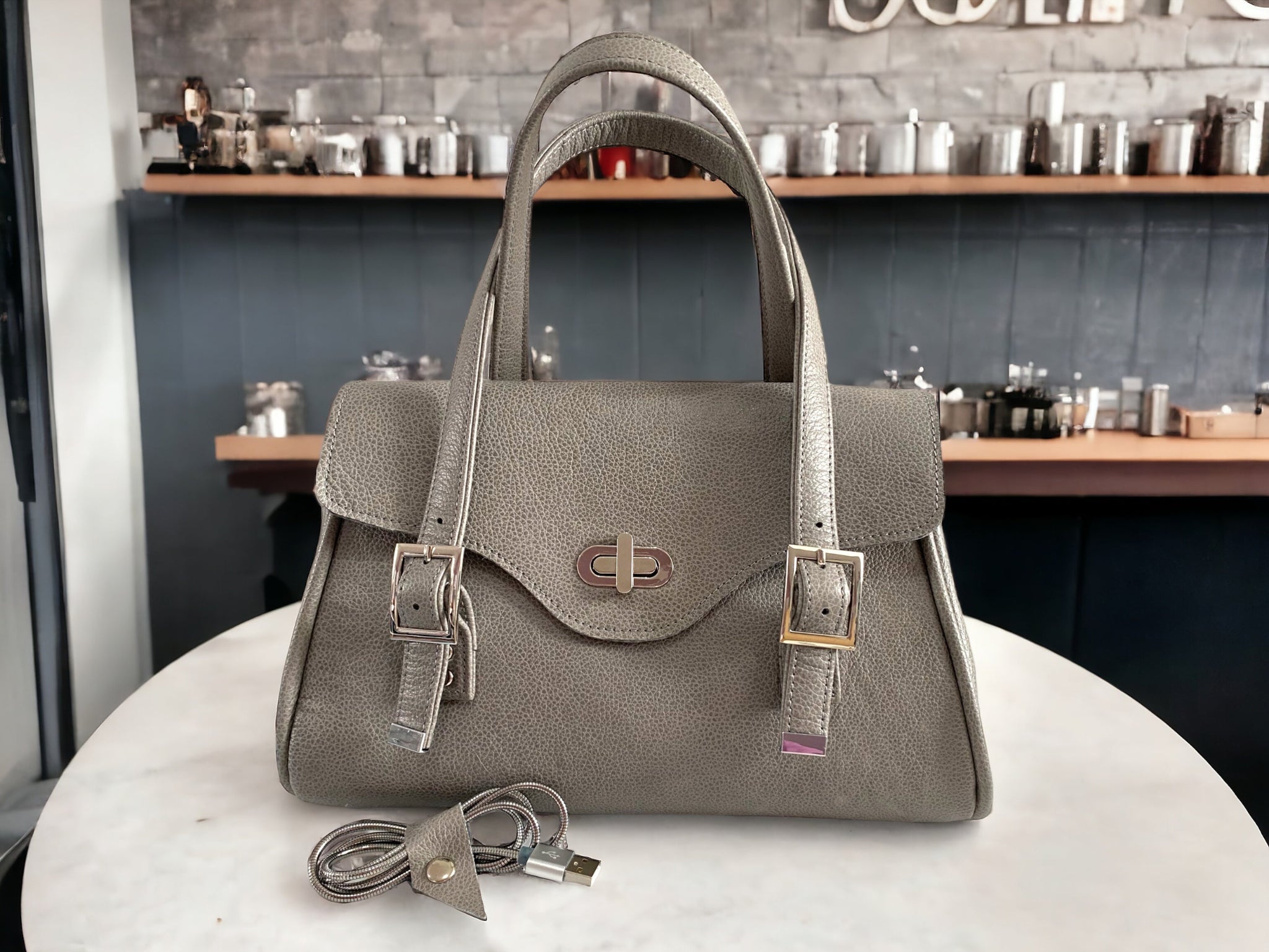 Grey leather handbag with silver hardware on a marble surface with a blurred cafe background.