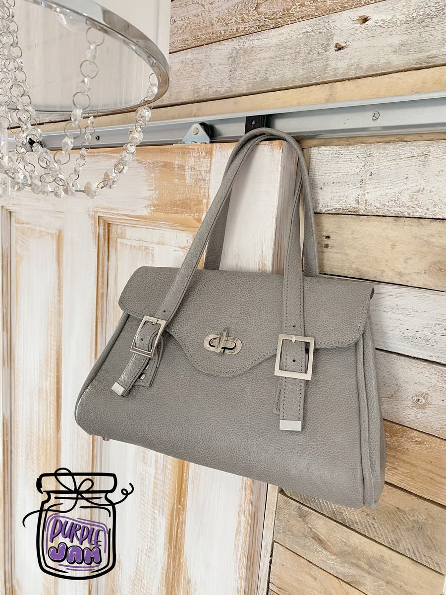 Grey leather handbag hanging on a wooden wall with a chandelier.