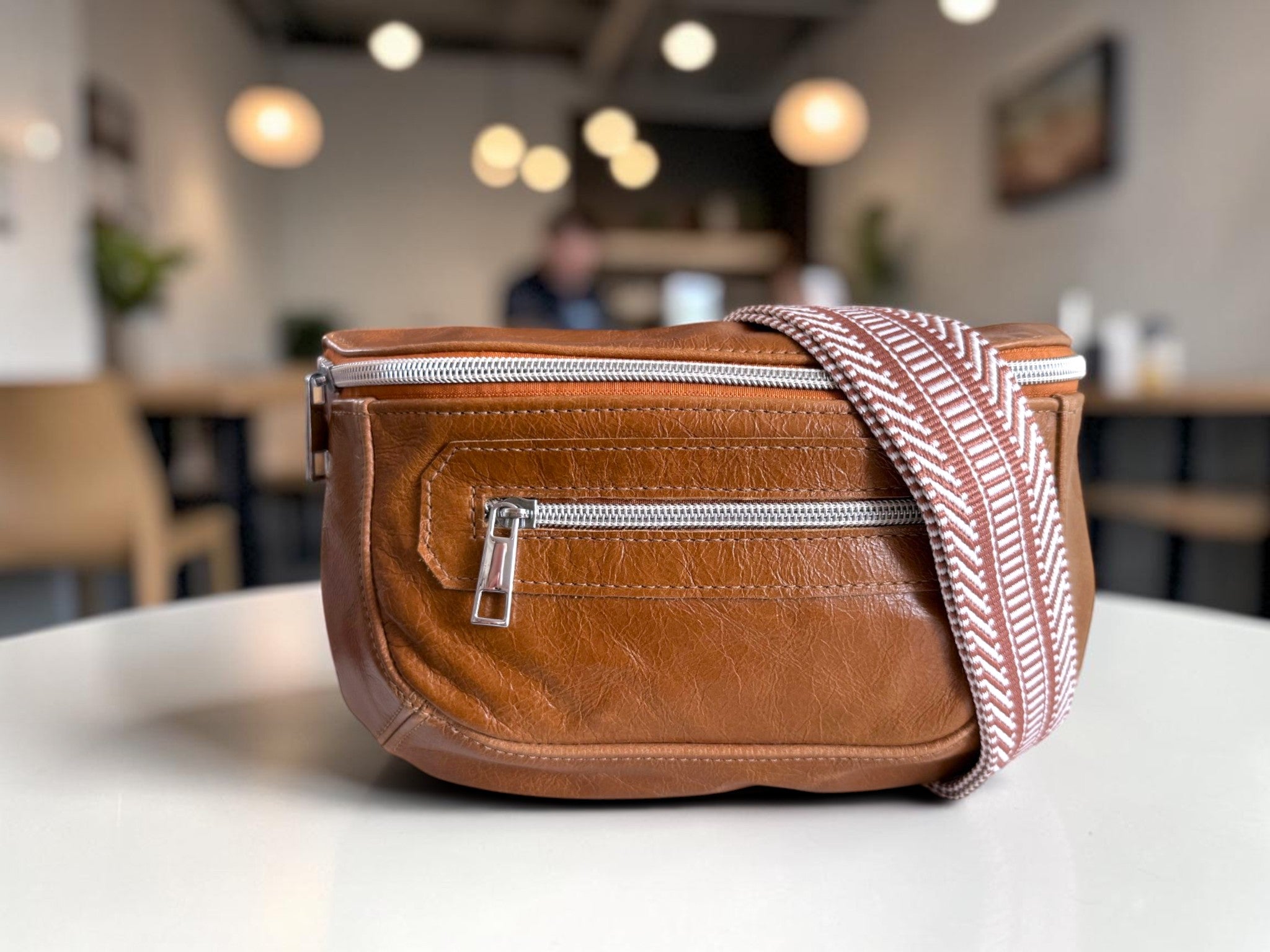 Tan brown leather sling bag with a patterned strap on a table in a blurred coffee shop setting