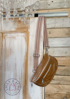 Tan brown leather bag with a strap hanging on a wooden door, with a chandelier in the background.