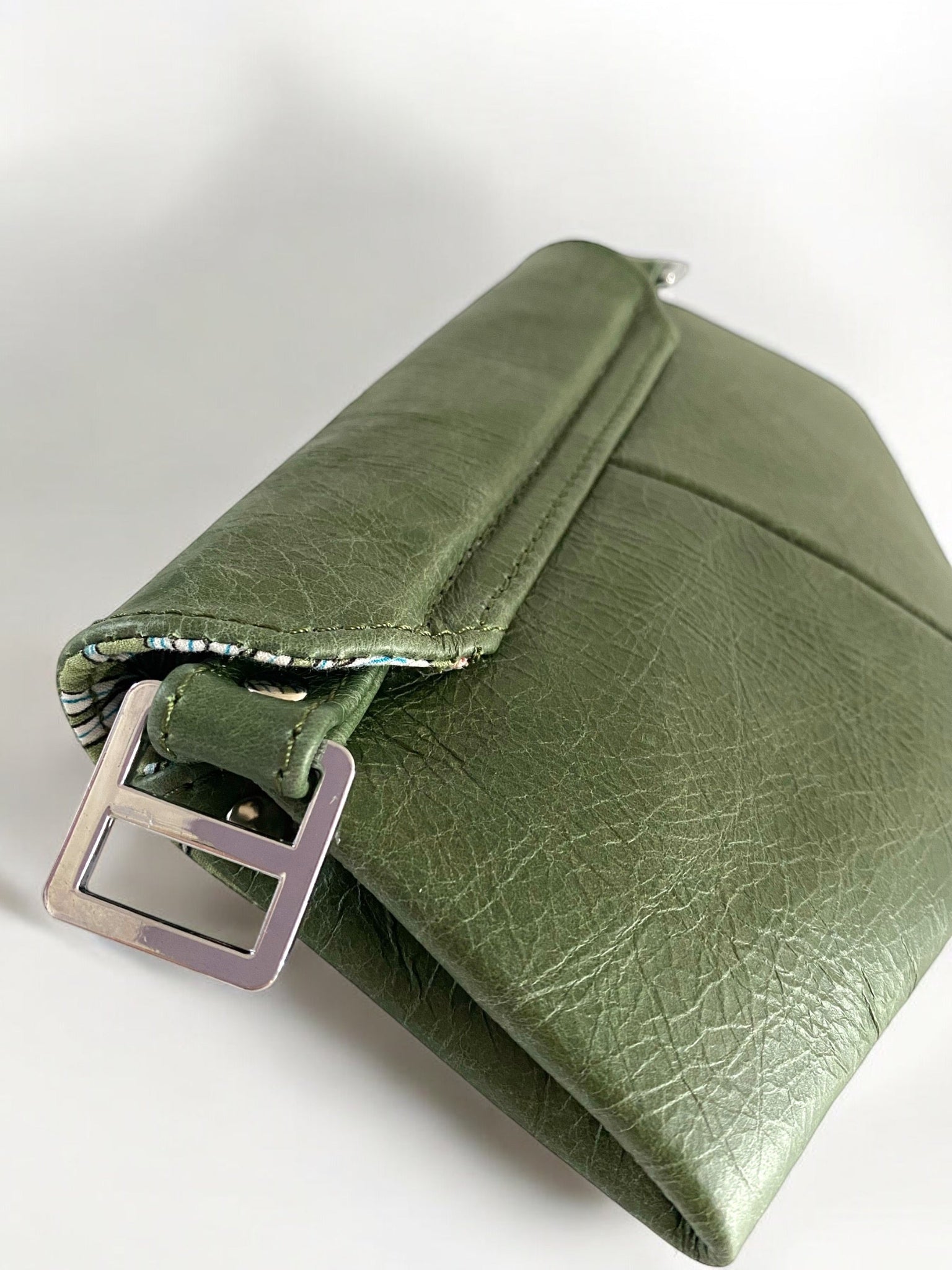 View of the back of a green, real leather clutch with metal buckle on white background