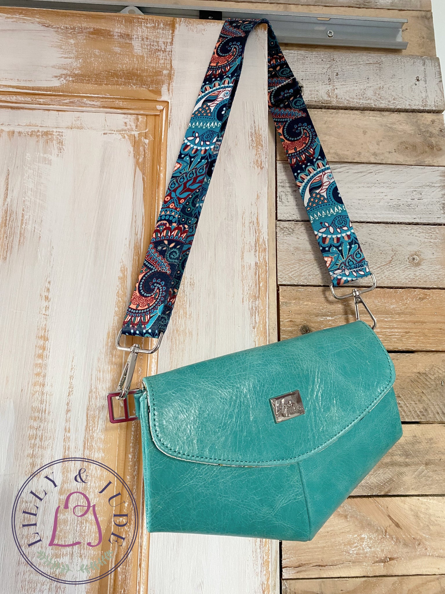 Teal leather crossbody bag with colorful paisley patterned strap on wooden door, with a Lilly & Jude brand logo