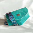 A turquoise leather crossbody clutch bag with a vibrant paisley patterned strap and a silver buckle.