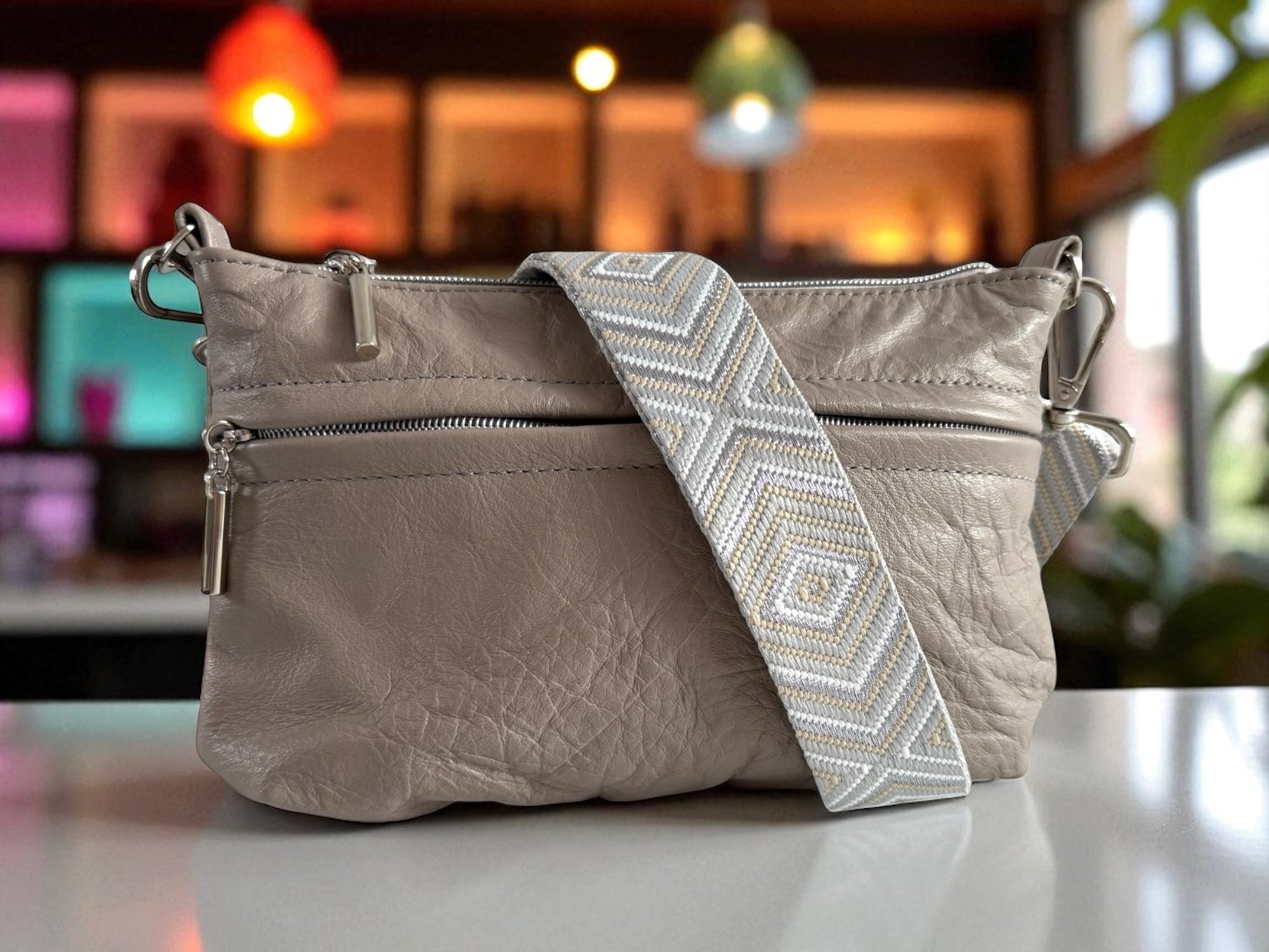 Taupe grey crossbody bag on café table with blurred colourful background, everyday leather bag by UK brand Lilly & Jude.

