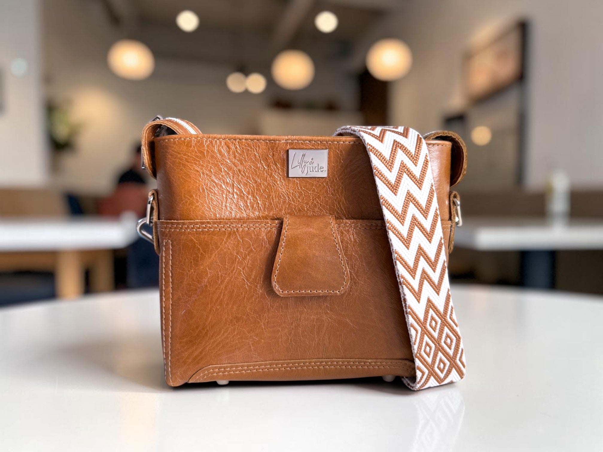 Tan brown leather crossbody bag with a patterned strap on a white surface, blurred coffee shop background